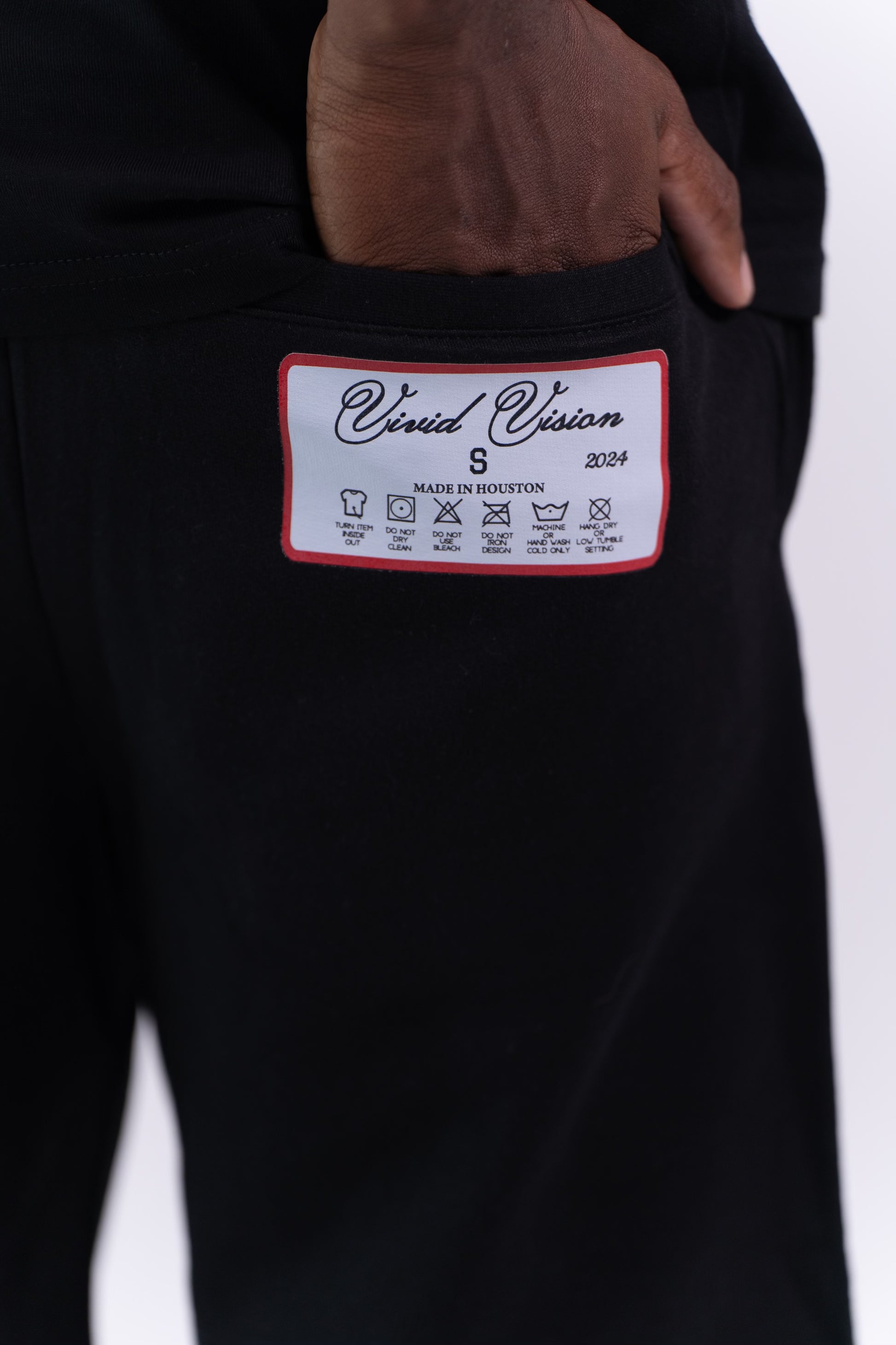 Someone wearing Vivid Vision Signature Shorts in black cotton, featuring a Vivid signature logo in large silver script on the side. They pair them with a black shirt and white socks against a plain, light background, revealing only their lower torso and legs for a stylishly practical look.
