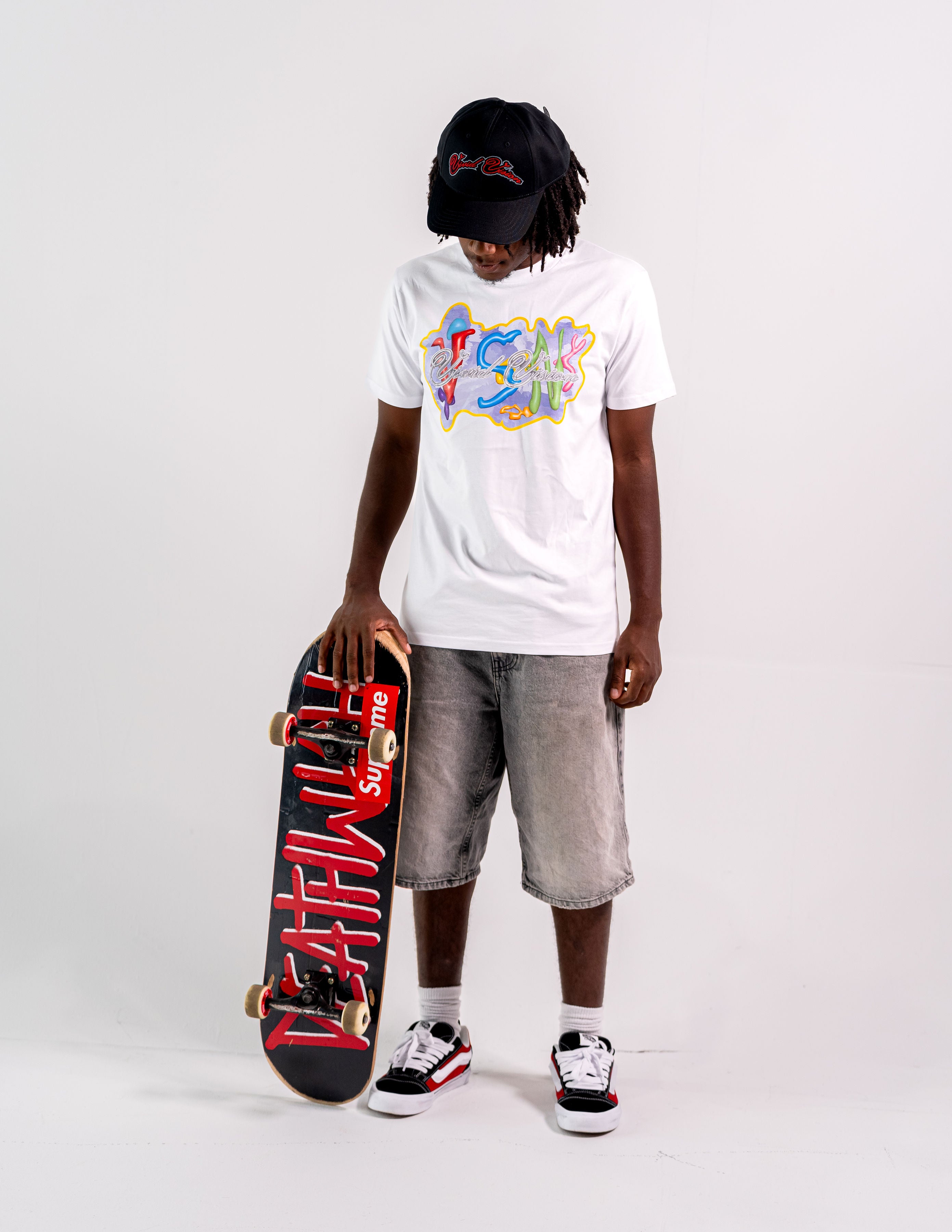 A person wearing a white t-shirt, denim shorts, and a black cap stands holding a skateboard with a red deck and white wheels. The skateboard features a bold design in white. They wear black and white sneakers. The background is plain white.