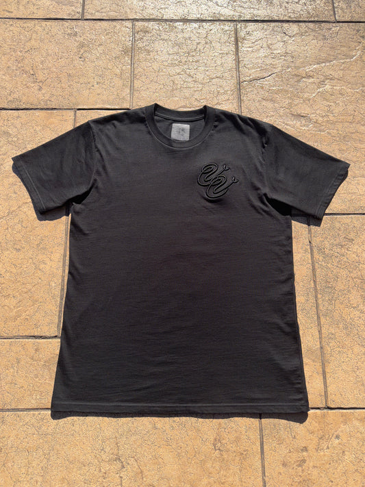 The Vivid Vision Black Embroidered Logo T-Shirt is laid flat on a textured tan stone surface, featuring a small black intertwined snakes design on the left chest and a visible garment tag inside the collar.