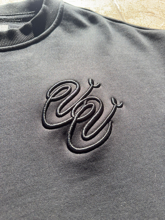The Vivid Vision Black Embroidered Logo Sweater is laid flat on a tan textured tile floor in direct sunlight, with its small W logo on the left chest and defined shadows at the edges.