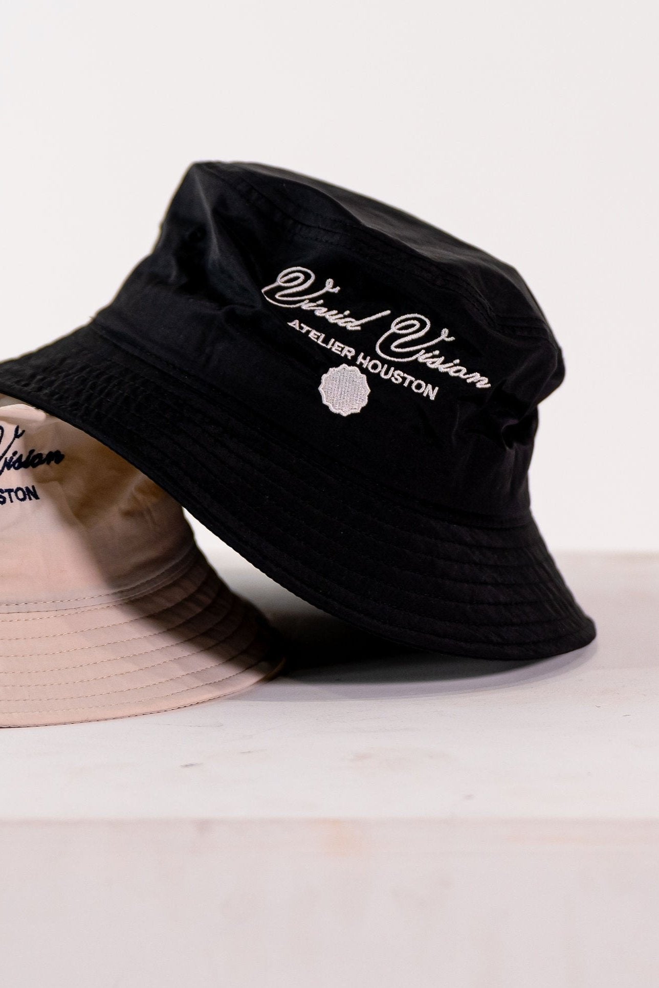 Two bucket hats are displayed on a white surface. One hat is black with white embroidered text reading Vivid Vision Atelier Houston. The other hat is beige and partially visible. Both hats have a simple, casual design.