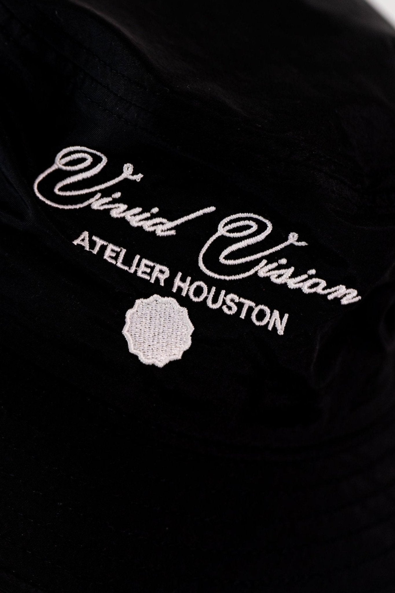 A close up of a black bucket hat with white embroidered text reading Vivid Vision Atelier Houston.