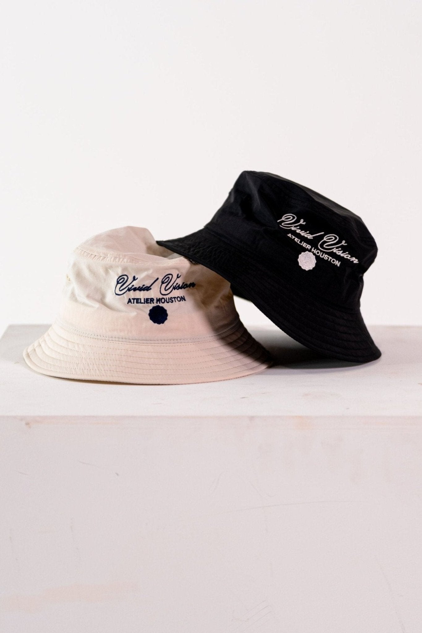 Two bucket hats are displayed on a white surface. One hat is black with white embroidered text reading Vivid Vision Atelier Houston. The other hat is beige and partially visible. Both hats have a simple, casual design.