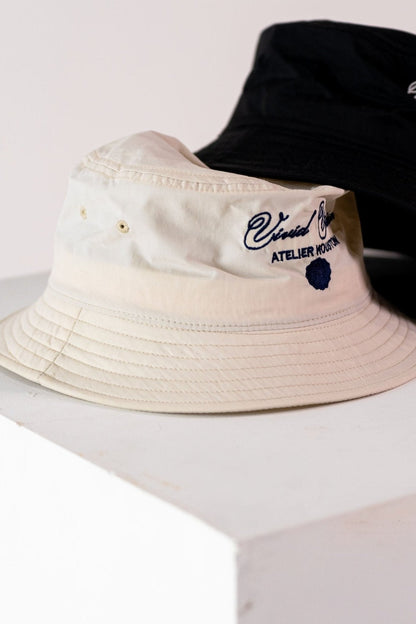 Two bucket hats are displayed on a white surface. One hat is beige with white embroidered text reading Vivid Vision Atelier Houston. The other hat is black and partially visible. Both hats have a simple, casual design.