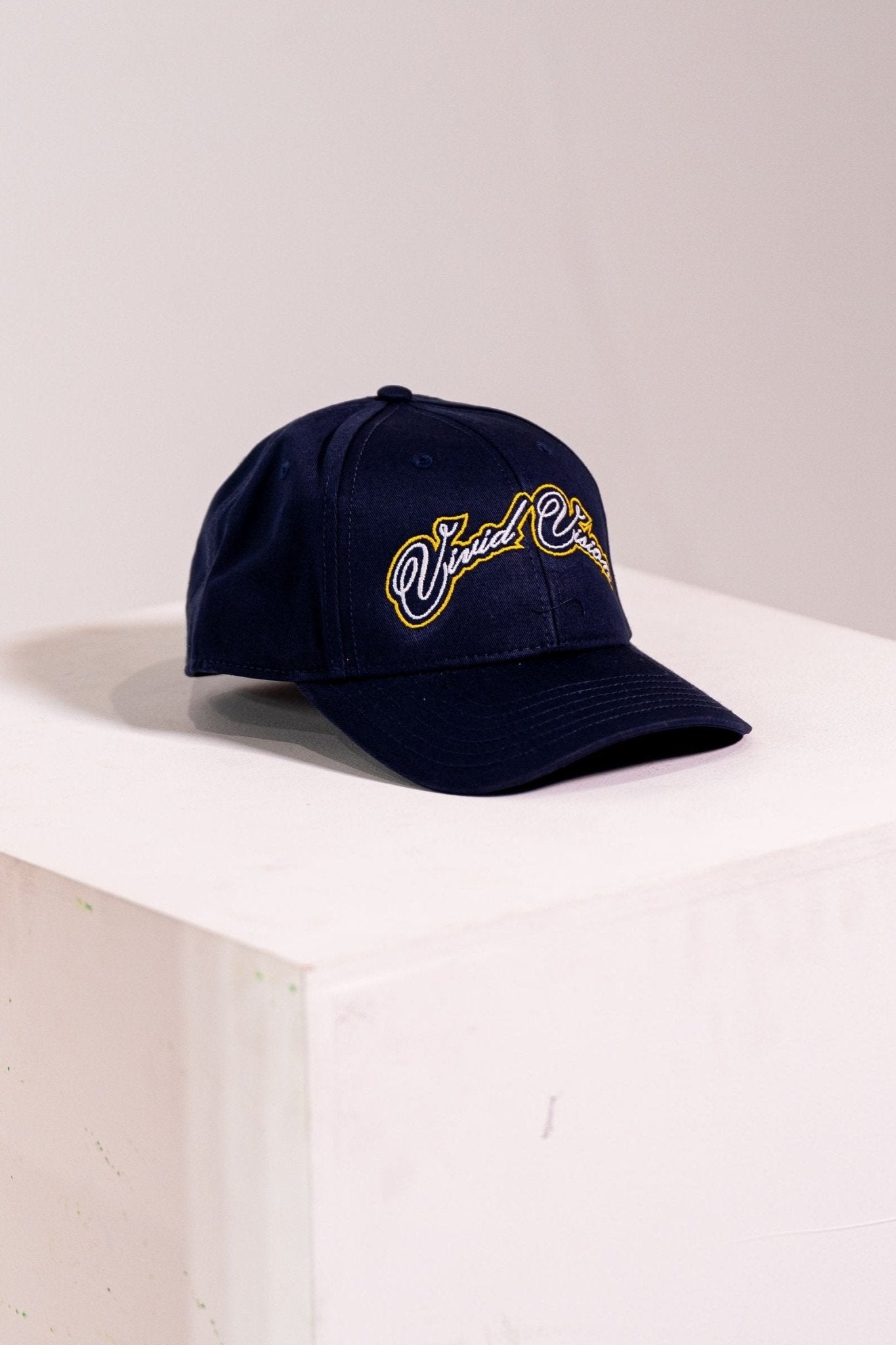 A navy baseball cap with white and yellow, cursive embroidery on the front. The cap is displayed on a white, square pedestal against a plain background.