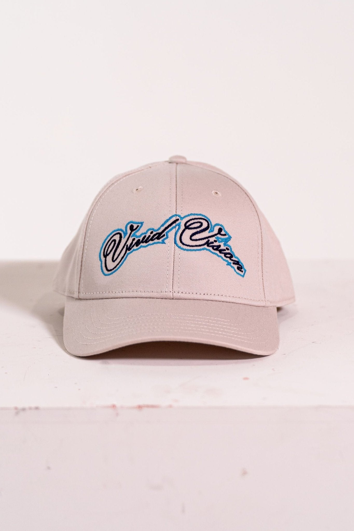 A beige baseball cap with light and navy blue, cursive embroidery on the front. The cap is displayed on a white, square pedestal against a plain background.