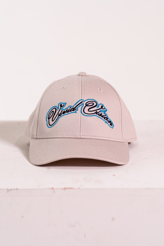 A beige baseball cap with light and navy blue, cursive embroidery on the front. The cap is displayed on a white, square pedestal against a plain background.