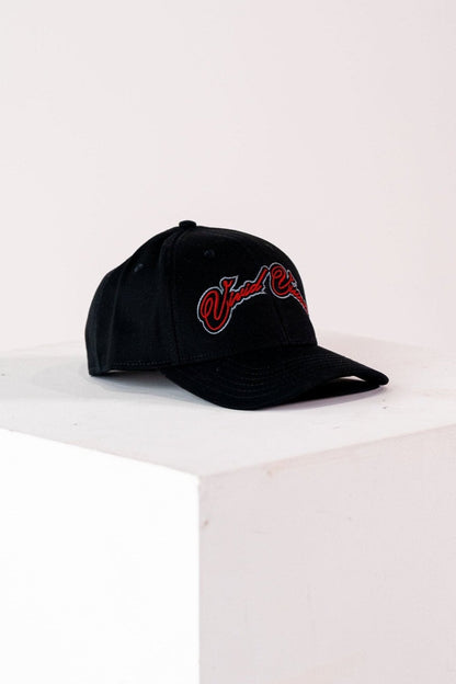 A black baseball cap with red and grey, cursive embroidery on the front. The cap is displayed on a white, square pedestal against a plain background.