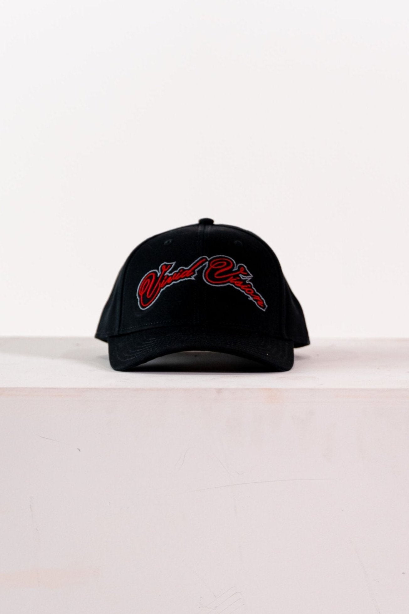 A black baseball cap with red and grey, cursive embroidery on the front. The cap is displayed on a white, square pedestal against a plain background.