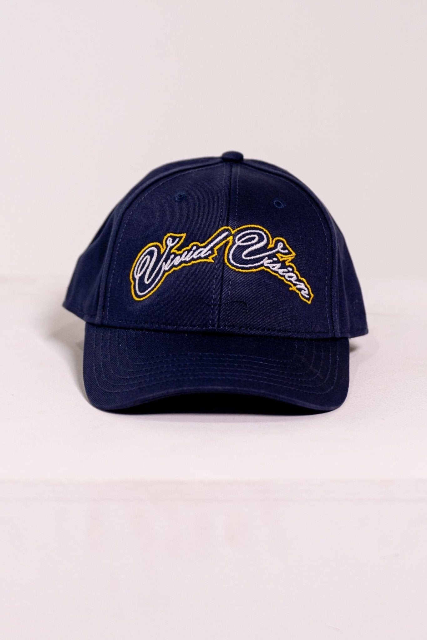 A navy baseball cap with white and yellow, cursive embroidery on the front. The cap is displayed on a white, square pedestal against a plain background.