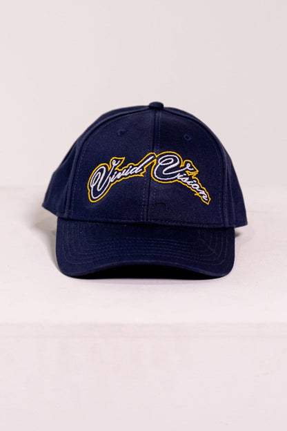 A navy baseball cap with white and yellow, cursive embroidery on the front. The cap is displayed on a white, square pedestal against a plain background.