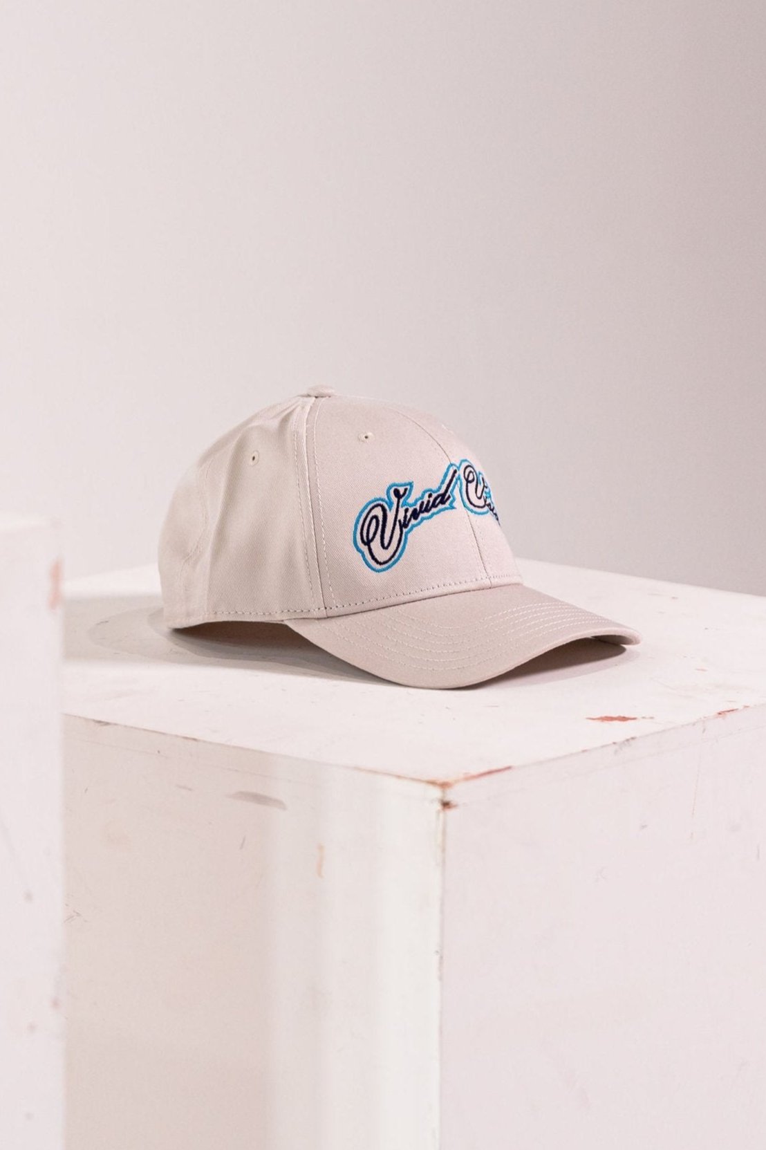 A beige baseball cap with light and navy blue, cursive embroidery on the front. The cap is displayed on a white, square pedestal against a plain background.